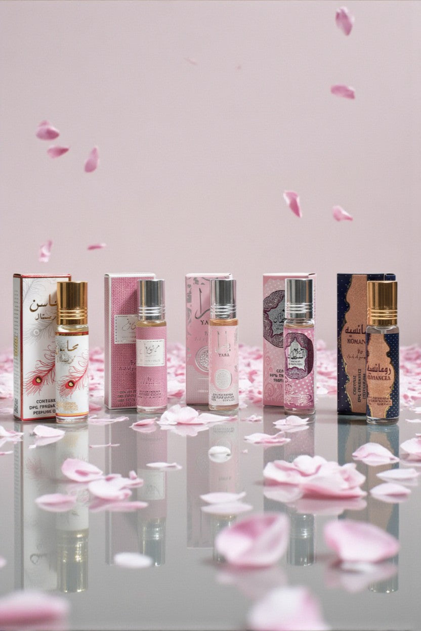 Pack 5 roll-on sans alcool Ard Al Zaafaran 10 ml – Mahasin Crystal, Mousuf Wardi, Yara, Hareem Al Sultan, Romancea, flacons alignés devant leurs étuis roses, reflet sur surface brillante avec pétales de fleurs.